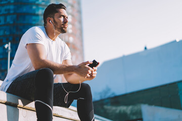 Pensive male bodybuilder in sportswear sitting at urbanity pondering on morning workout exercises, Caucasian athlete with cellphone gadget in hands listening audio podcast during training break