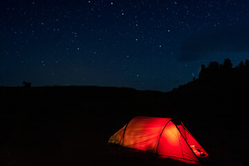 Glowing red tent with hikers sleeping in the wild mountains under dramatic night sky. Lit camping tent in the forest under starry sky during summer vacation.