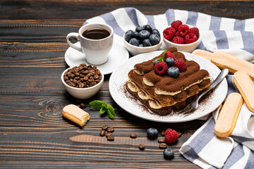 portion of Classic tiramisu dessert with raspberries and blueberries, savoiardi cookies and cup of espresso coffee isolated on wooden background