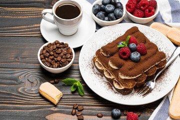 portion of Classic tiramisu dessert with raspberries and blueberries, savoiardi cookies and cup of espresso coffee isolated on wooden background