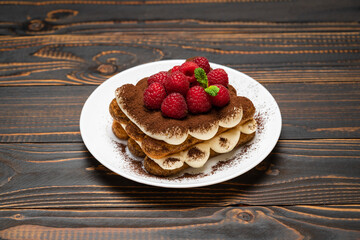portion of Classic tiramisu dessert with raspberries on ceramic plate on wooden background