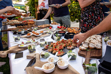 Outdoor garden party with buffet table full of canapes. People serving themselves. Close up of hands taking finger food in their plates.  - obrazy, fototapety, plakaty