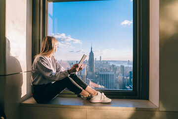 Thoughtful female freelancer with modern touch pad in hands looking in panoramic window with beautiful scenery of New York megalopolis, millennial woman with digital tablet dreaming about travel
