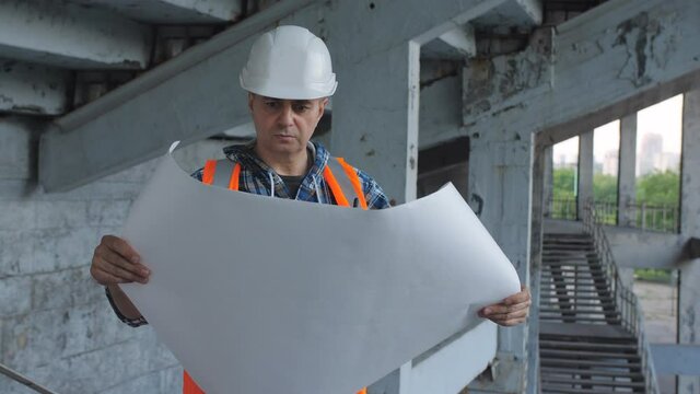A male Builder unfolds and examines the drawing of a building on the construction site.