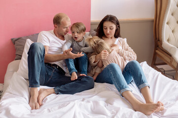 Beautiful young family: mom, dad, daughter and son are sitting on the bed at home in a bright bedroom and hugging
