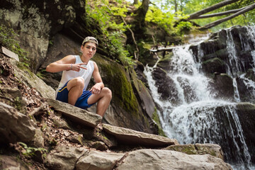 Next to a beautiful waterfall sits a teenage boy and enjoys nature, relaxing in the mountains, gaining strength for the hustle and bustle of the city and college