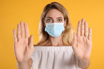 Woman in protective face mask showing stop gesture on yellow background. Prevent spreading of coronavirus