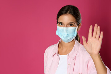 Woman in protective mask showing hello gesture on pink background, space for text. Keeping social distance during coronavirus pandemic