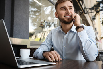 Happy young man with laptop talking on phone at outdoor cafe