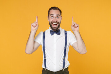 Fototapeta premium Cheerful excited young bearded man 20s in white shirt bow-tie suspender posing standing pointing index fingers up on mock up copy space isolated on bright yellow color wall background studio portrait.