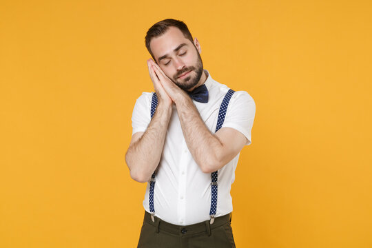 Relaxed young bearded man 20s in white shirt bow-tie suspender posing standing sleep with folded hands under cheek keeping eyes closed isolated on bright yellow color wall background studio portrait.