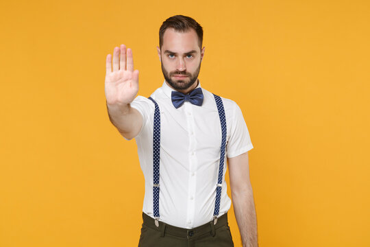 Displeased serious young bearded man 20s wearing white shirt bow-tie suspender posing showing stop gesture with palm looking camera isolated on bright yellow color wall background studio portrait.
