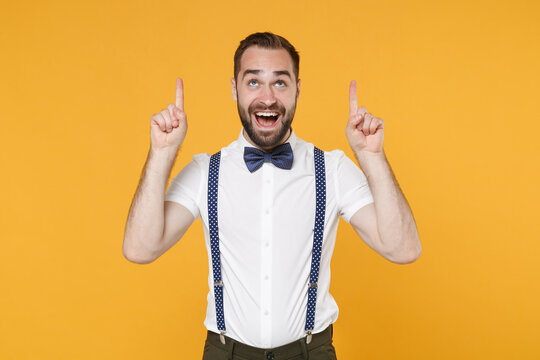 Cheerful excited young bearded man 20s in white shirt bow-tie suspender posing standing pointing index fingers up on mock up copy space isolated on bright yellow color wall background studio portrait.