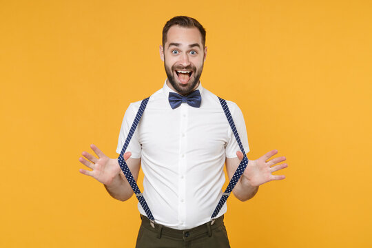 Excited young bearded man 20s wearing white shirt bow-tie posing standing stretching suspender showing making stick tongue out sign, fooling around isolated on yellow color background studio portrait.