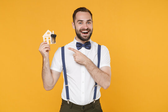 Funny young bearded man 20s wearing white shirt bow-tie suspender posing standing hold in hand pointing index finger on house bunch of keys isolated on bright yellow color background studio portrait.