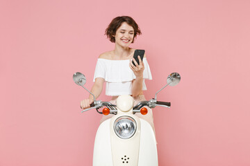Smiling beautiful young brunette woman 20s wearing white summer clothes using mobile cell phone typing sms message sitting driving moped isolated on pastel pink colour background, studio portrait.