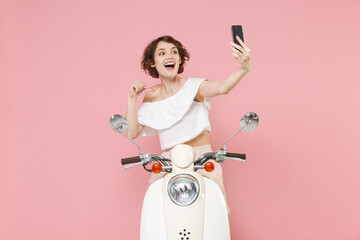 Excited young brunette woman 20s in white summer clothes posing doing selfie shot on mobile phone pointing thumb on herself driving moped isolated on pastel pink colour background, studio portrait.