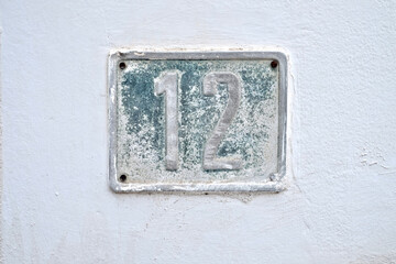 Number 12, twelve, on weathered blue plate on a white wall.