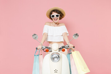 Cheerful young woman 20s wearing white summer clothes hat glasses hold package bag with purchases after shopping looking camera driving moped isolated on pastel pink colour background studio portrait.