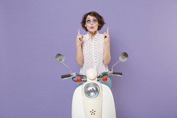 Shocked amazed young brunette woman in white dotted shirt glasses pointing index fingers up on mock up copy space sitting and driving moped isolated on pastel violet colour background studio portrait.