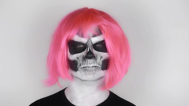 Cheerful funny man in halloween skeleton makeup in pink wig blowing hair off his face. Shooting in the studio. Gray background
