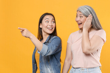 Excited family asian women girls gray-haired mother and brunette daughter in casual clothes posing pointing index finger aside put hand on cheek isolated on yellow color background studio portrait.