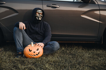 An unrecognizable adult man wearing a skull mask sits near a car on the grass and holds a carved...