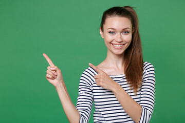 Smiling beautiful young brunette woman 20s wearing striped casual clothes posing standing pointing index fingers aside up on mock up copy space isolated on green color wall background studio portrait.
