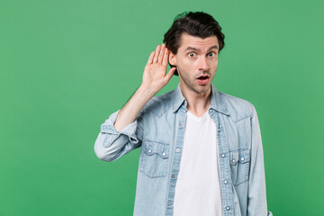 Shocked curious young brunet man 20s wearing casual clothes white t-shirt denim shirt posing try to hear you overhear listening intently looking camera isolated on green background studio portrait.