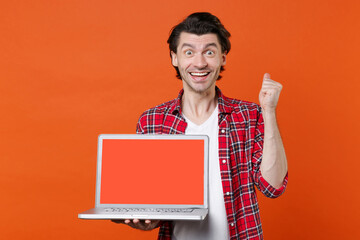 Excited young man 20s in white t-shirt red checkered shirt posing hold laptop pc computer with blank empty screen mock up copy space doing winner gesture isolated on orange background studio portrait.