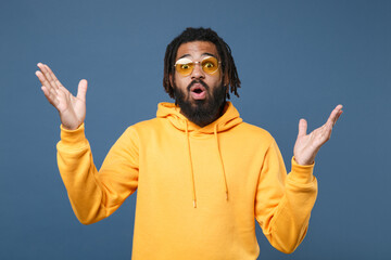 Shocked young african american man guy in yellow streetwear hoodie glasses isolated on blue...