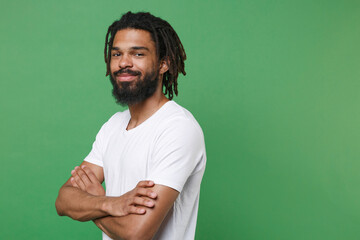 Side view of smiling funny young african american man guy 20s wearing white casual t-shirt posing holding hands folded crossed looking camera isolated on green color wall background studio portrait.