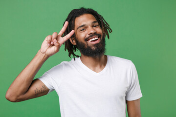 Fototapeta premium Smiling funny cheerful young african american man guy 20s wearing white casual t-shirt posing showing victory sign looking camera isolated on green color wall background studio portrait.