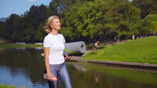 Active senior woman with fitness mat and water bottle in hands walking in park