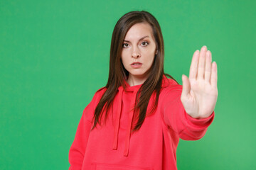 Serious concerned young brunette woman 20s wearing bright red casual streetwear hoodie posing showing stop gesture with palm looking camera isolated on green color wall background studio portrait.