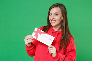 Smiling beautiful young brunette woman 20s wearing bright red casual streetwear hoodie posing holding in hands gift certificate looking camera isolated on green color wall background studio portrait.