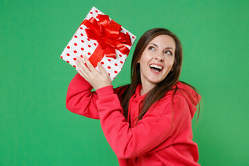 Cheerful curious funny young brunette woman in red casual streetwear hoodie posing celebrating hold red present box with gift ribbon bow looking up isolated on green color background studio portrait.