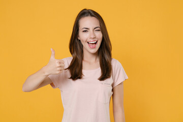 Cheerful funny blinking young brunette woman 20s wearing pastel pink casual t-shirt posing standing showing thumb up looking camera isolated on bright yellow color wall background studio portrait.