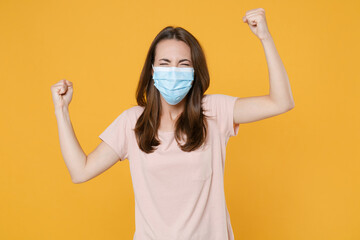 Happy young brunette woman in pastel pink casual t-shirt sterile face mask to safe from coronavirus virus covid-19 2019-ncov doing winner gesture isolated on yellow color background studio portrait.