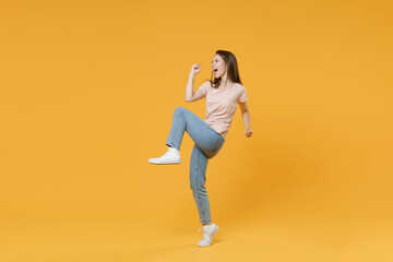 Full length side view portrait of happy joyful young woman wearing pastel pink casual t-shirt posing clenching fists doing winner gesture rising leg isolated on yellow color wall background studio.