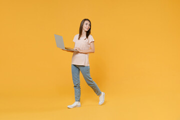 Full length portrait of smiling young brunette woman 20s wearing pastel pink casual t-shirt posing working on laptop pc computer looking aside isolated on bright yellow color wall background studio.