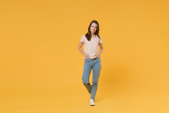 Full length portrait of smiling beautiful charming young woman 20s in pastel pink casual t-shirt posing holding hands in pockets looking camera isolated on bright yellow color wall background studio.