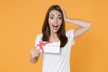 Excited surprised young brunette woman 20s wearing white blank casual t-shirt posing hold gift certificate keeping mouth open put hand on head isolated on yellow color wall background studio portrait.