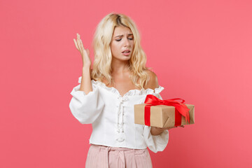 Displeased young woman in white casual clothes hold red present box with gift ribbon bow spreading hands isolated on pink color background studio. Valentine's Day Women's Day birthday holiday concept.