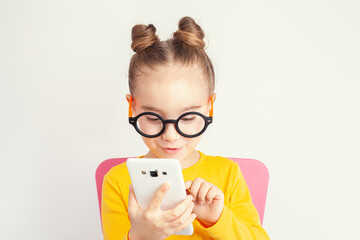 Beautiful cute little girl with eyeglasses holding smartphone in her hand and making gesture on screen. Child from alpha generation playing or using mobile app or playing game on smartphone. .