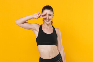 Fototapeta na wymiar Smiling blinking young fitness sporty woman 20s wearing black sportswear posing training working out showing victory sign looking camera isolated on bright yellow color background studio portrait.