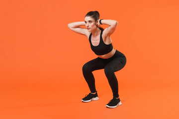 Fototapeta na wymiar Full length portrait of strong young fitness sporty woman in black sportswear training working out doing exercise squatting with hands behind head looking aside isolated on orange background studio.