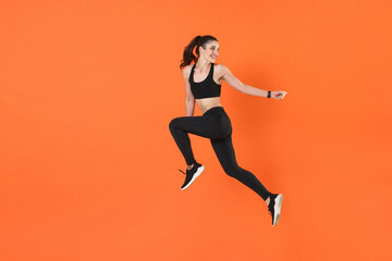 Fototapeta na wymiar Full length side view of portrait of funny young fitness sporty woman 20s in black sportswear posing training working out jumping like running looking aside isolated on orange color background studio.