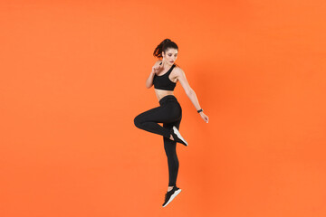 Full length side view of portrait of young fitness sporty woman 20s wearing black sportswear posing training working out jumping like running looking aside isolated on orange color background studio.