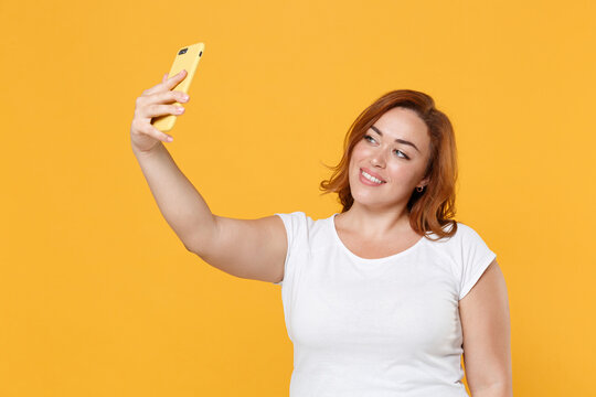 Smiling beautiful young redhead plus size body positive female woman girl 20s in white casual t-shirt posing doing selfie shot on mobile phone isolated on yellow color wall background studio portrait.